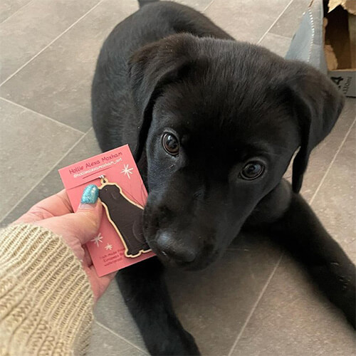 A hand holding a wooden black Labrador keyring on a pink backing card in front of a black Labrador puppy.