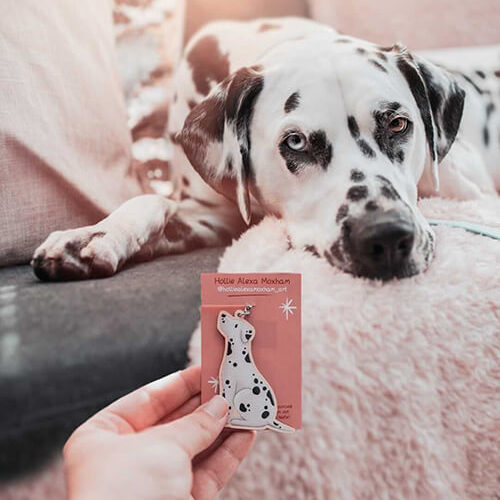 A hand holding a wooden Dalmatian keyring on a pink backing card in front of a Dalmatian dog.
